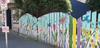 A vibrant wooden fence is adorned with colorful floral and nature-themed paintings, featuring flowers, butterflies, and abstract patterns. The fence panels vary in color, including shades of blue, green, yellow, and purple. A 'No Parking' sign is visible nearby, along with trees providing shade.
