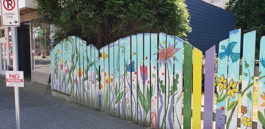 A vibrant wooden fence is adorned with colorful floral and nature-themed paintings, featuring flowers, butterflies, and abstract patterns. The fence panels vary in color, including shades of blue, green, yellow, and purple. A 'No Parking' sign is visible nearby, along with trees providing shade.