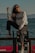 A person wearing sunglasses, a white sweatshirt with the word 'ATELIER' printed on it, and dark ripped jeans is posing by the seaside. The individual is sitting with one leg raised on a rail that is decorated with various stickers. There is a 'No Diving' sign at the bottom of the rail. The background features a calm sea and distant mountains under a clear sky.