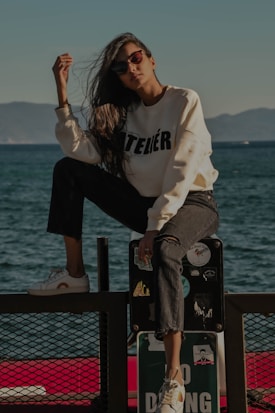 A person wearing sunglasses, a white sweatshirt with the word 'ATELIER' printed on it, and dark ripped jeans is posing by the seaside. The individual is sitting with one leg raised on a rail that is decorated with various stickers. There is a 'No Diving' sign at the bottom of the rail. The background features a calm sea and distant mountains under a clear sky.