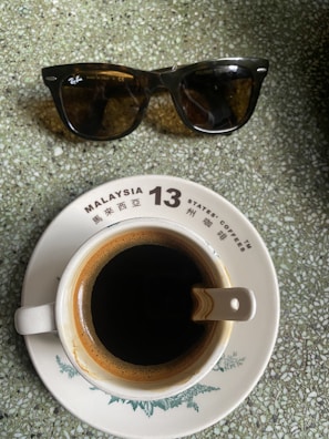 A pair of sunglasses sits above a ceramic cup of black coffee on a saucer. The saucer contains text indicating 'Malaysia 13 States Coffees' in both English and Chinese. The surface beneath them has a textured, speckled green appearance.