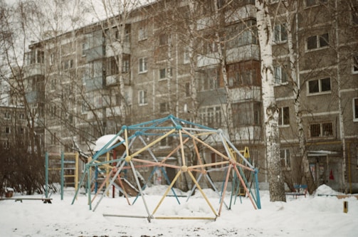A snowy playground with a geometric climbing structure made of metal rods painted in various colors, surrounded by leafless birch trees and apartment buildings in the background.