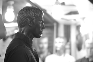 A black and white photograph features a close-up side profile of a mannequin with stylized hair and a collared shirt. In the background, blurred outlines of other mannequins and hanging light bulbs are visible, giving a surreal and artistic atmosphere.
