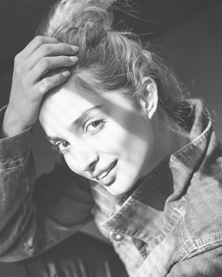 A woman with light hair is shown in a high-contrast black and white photograph. She is touching her hair with one hand and wearing a denim jacket. The image captures a close-up of her smiling face, highlighted by soft shadows and bright light.