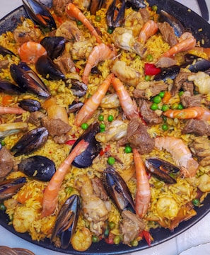A large skillet filled with a colorful and appetizing dish called paella, featuring a mix of vibrant yellow rice, plump shrimp, mussels, peas, and various cuts of meat. The shrimp are large and orange, contrasting with the dark shells of the mussels. The rice is interspersed with green peas and chunks of vegetables, adding to the dish's visual appeal.