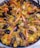 A large skillet filled with a colorful and appetizing dish called paella, featuring a mix of vibrant yellow rice, plump shrimp, mussels, peas, and various cuts of meat. The shrimp are large and orange, contrasting with the dark shells of the mussels. The rice is interspersed with green peas and chunks of vegetables, adding to the dish's visual appeal.