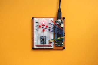 A breadboard with electronic components, including resistors and colorful jumper wires, is connected to a microcontroller on a wooden base. The setup is laid out on a vibrant yellow background.