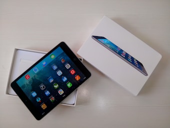 A tablet is displayed partially out of its open white box, which features an image of the tablet on its cover. The screen of the tablet shows a variety of colorful app icons against a galaxy-themed background. Both the device and the packaging are placed on a light-colored surface.