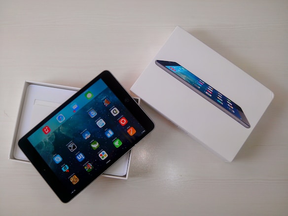 A tablet is displayed partially out of its open white box, which features an image of the tablet on its cover. The screen of the tablet shows a variety of colorful app icons against a galaxy-themed background. Both the device and the packaging are placed on a light-colored surface.