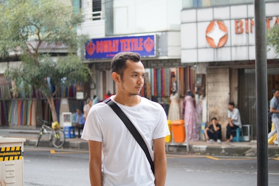 A man is standing on a city street wearing a white t-shirt with a black shoulder strap. Behind him, there are shops displaying colorful textiles and mannequins with dresses, while people are lounging around the area. There are trees and a bicycle visible, contributing to a busy urban scene.