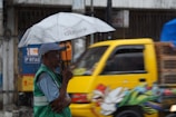 A person stands under a grey umbrella with a 'Citibank' logo, wearing a green vest and a cap. They are positioned on a street while it is raining, and a yellow vehicle decorated with colorful artwork drives by in the background.