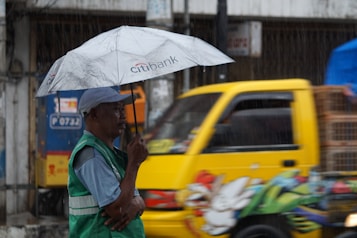 A person stands under a grey umbrella with a 'Citibank' logo, wearing a green vest and a cap. They are positioned on a street while it is raining, and a yellow vehicle decorated with colorful artwork drives by in the background.