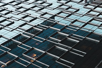 An abstract architectural pattern created by a glass building facade with protruding geometric window frames. The reflective surfaces display a grid-like arrangement with a mix of angles and shadows, suggesting depth and complexity.
