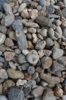 A detailed array of pebbles and stones with varied textures and colors, including grey, beige, and muted earth tones, scattered closely together. The stones vary in size and shape, creating a natural mosaic effect.