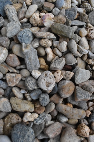 A detailed array of pebbles and stones with varied textures and colors, including grey, beige, and muted earth tones, scattered closely together. The stones vary in size and shape, creating a natural mosaic effect.