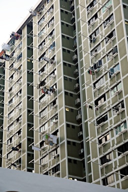 A high-rise residential building features numerous windows and balconies, with multiple clothes drying on rods extending from the windows. The facade consists of muted green panels with white framing creating a grid-like pattern.