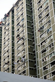 A high-rise residential building features numerous windows and balconies, with multiple clothes drying on rods extending from the windows. The facade consists of muted green panels with white framing creating a grid-like pattern.