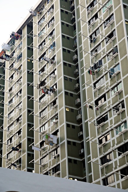 A high-rise residential building features numerous windows and balconies, with multiple clothes drying on rods extending from the windows. The facade consists of muted green panels with white framing creating a grid-like pattern.