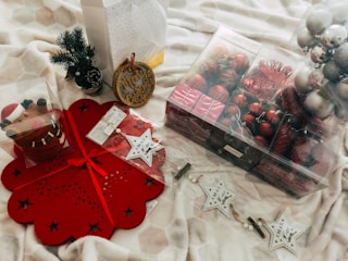 A collection of Christmas decorations is spread out on a beige patterned textile. There are red and silver baubles in a transparent box, a miniature pine tree, and a festive candle holder featuring a reindeer design. A red snowflake-shaped placemat and white star-shaped ornaments with the word 'XMAS' are also visible.