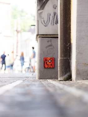 A small, vibrant piece of urban art depicting a face with bold expression is attached to a concrete column in an alleyway. The foreground features a graffiti-covered surface, while the background shows several pedestrians walking, slightly out of focus.