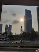 A cityscape featuring a tall, uniquely-shaped skyscraper under a partly cloudy sky with the sun visible. Surrounding the skyscraper are other tall buildings, trees, and a busy road with cars. Urban infrastructure such as streetlights and communication antennas are noticeable.