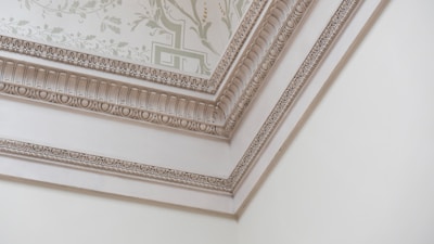 A corner of a room featuring intricate decorative molding on the ceiling and walls. The molding includes ornate patterns and floral designs, with soft, neutral colors enhancing the elegance. The ceiling has a light-colored wallpaper with a subtle floral motif.