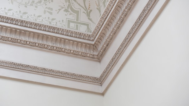 A corner of a room featuring intricate decorative molding on the ceiling and walls. The molding includes ornate patterns and floral designs, with soft, neutral colors enhancing the elegance. The ceiling has a light-colored wallpaper with a subtle floral motif.