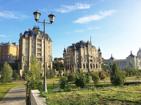Elegant historic buildings with ornate architectural details are surrounded by well-maintained green gardens. A clear blue sky enhances the charm of the location, with towering trees and neatly arranged pathways.