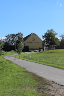 A paved pathway winds through a grassy area, leading to a large yellow building with a brown roof. Trees flank the pathway, and the words 'HŘEBČÍN RUF' are prominently displayed on the building along with a horse graphic.