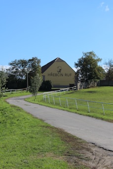 A paved pathway winds through a grassy area, leading to a large yellow building with a brown roof. Trees flank the pathway, and the words 'HŘEBČÍN RUF' are prominently displayed on the building along with a horse graphic.