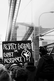 A black and white photo of a protest scene with a prominent sign that reads 'Respect Existence or Expect Resistance' alongside an image of a raised fist. The background includes reflective surfaces of buildings and a crowd of people.