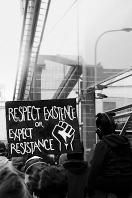 A black and white photo of a protest scene with a prominent sign that reads 'Respect Existence or Expect Resistance' alongside an image of a raised fist. The background includes reflective surfaces of buildings and a crowd of people.