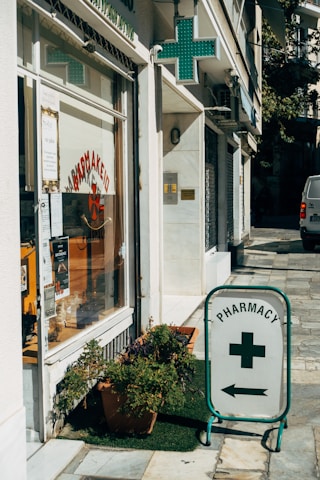 A street corner features a pharmacy with a glass storefront displaying signs in Greek and various products. There is a prominent pharmacy sign featuring a green cross and an arrow pointing towards the store entrance. Potted plants are placed on the sidewalk, adding a touch of greenery to the urban setting.