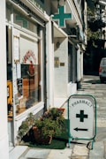 A street corner features a pharmacy with a glass storefront displaying signs in Greek and various products. There is a prominent pharmacy sign featuring a green cross and an arrow pointing towards the store entrance. Potted plants are placed on the sidewalk, adding a touch of greenery to the urban setting.
