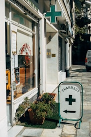 A street corner features a pharmacy with a glass storefront displaying signs in Greek and various products. There is a prominent pharmacy sign featuring a green cross and an arrow pointing towards the store entrance. Potted plants are placed on the sidewalk, adding a touch of greenery to the urban setting.