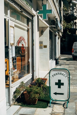 A street corner features a pharmacy with a glass storefront displaying signs in Greek and various products. There is a prominent pharmacy sign featuring a green cross and an arrow pointing towards the store entrance. Potted plants are placed on the sidewalk, adding a touch of greenery to the urban setting.