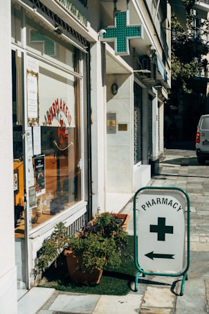 A street corner features a pharmacy with a glass storefront displaying signs in Greek and various products. There is a prominent pharmacy sign featuring a green cross and an arrow pointing towards the store entrance. Potted plants are placed on the sidewalk, adding a touch of greenery to the urban setting.