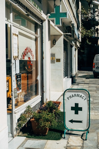 A street corner features a pharmacy with a glass storefront displaying signs in Greek and various products. There is a prominent pharmacy sign featuring a green cross and an arrow pointing towards the store entrance. Potted plants are placed on the sidewalk, adding a touch of greenery to the urban setting.