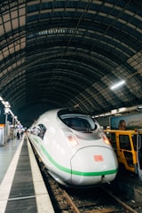A sleek, modern high-speed train is stationed on a railway track under a large, curved metal and glass structure. The train is predominantly white with green accents and has an aerodynamic design. The platform alongside the train is lined with lights, and there is a yellow maintenance vehicle parked nearby.