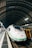 A sleek, modern high-speed train is stationed on a railway track under a large, curved metal and glass structure. The train is predominantly white with green accents and has an aerodynamic design. The platform alongside the train is lined with lights, and there is a yellow maintenance vehicle parked nearby.