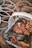 A white woven basket filled with autumn-themed items such as a fabric pumpkin, red berries, dried leaves, and a pinecone, resting on a textured brown blanket. The scene is accented by a white wire decorative frame.