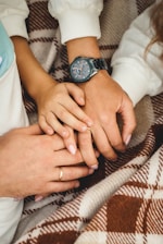 Several hands are gently resting on each other, symbolizing connection and affection. A wristwatch is visible on one of the wrists, and all the hands are placed on a plaid blanket.
