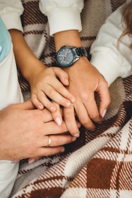Several hands are gently resting on each other, symbolizing connection and affection. A wristwatch is visible on one of the wrists, and all the hands are placed on a plaid blanket.