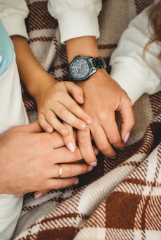Several hands are gently resting on each other, symbolizing connection and affection. A wristwatch is visible on one of the wrists, and all the hands are placed on a plaid blanket.