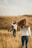 In a grassy field, a person with long hair walks towards another who is joyfully lifting and holding a child. The scene conveys a sense of motion and playfulness.