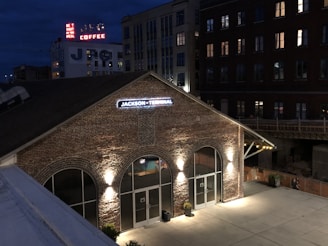 A brick building with a sign reading 'Jackson Terminal' is illuminated by exterior lights at night. The surrounding area includes other taller buildings, some with lit windows. A vintage-style neon sign advertising coffee and a message can be seen in the background.