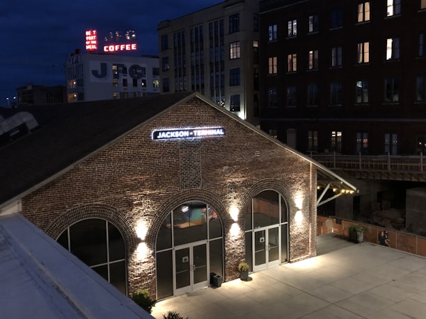 A brick building with a sign reading 'Jackson Terminal' is illuminated by exterior lights at night. The surrounding area includes other taller buildings, some with lit windows. A vintage-style neon sign advertising coffee and a message can be seen in the background.