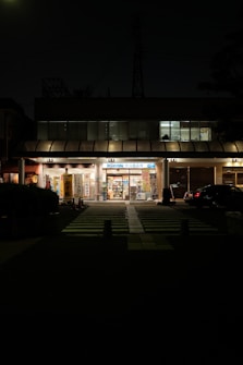 A small convenience store is illuminated in the darkness, with a bright entrance displaying various signs and posters. The building has a modern look with tall glass windows above. A crosswalk in the foreground leads to the store, and a car is parked nearby.