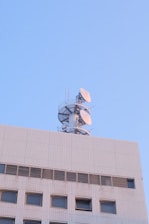 A building structure with a communication tower on top, featuring several satellite dishes against a clear blue sky.