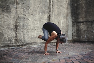 A person is performing a yoga pose, balancing on their hands with knees tucked close to the arms, against a textured stone wall. The individual has gray hair tied back and is wearing a black top and gray leggings. The setting appears to be outdoors on a brick platform.